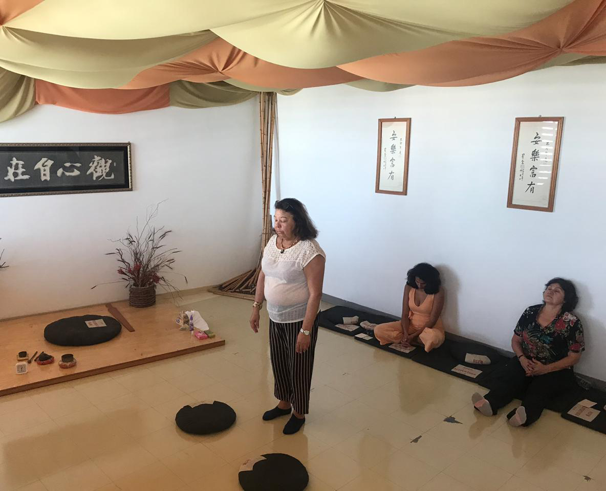 Imagem de uma mulher, em pé, meditando, enquanto outras duas estão meditando sentadas no lado direito.