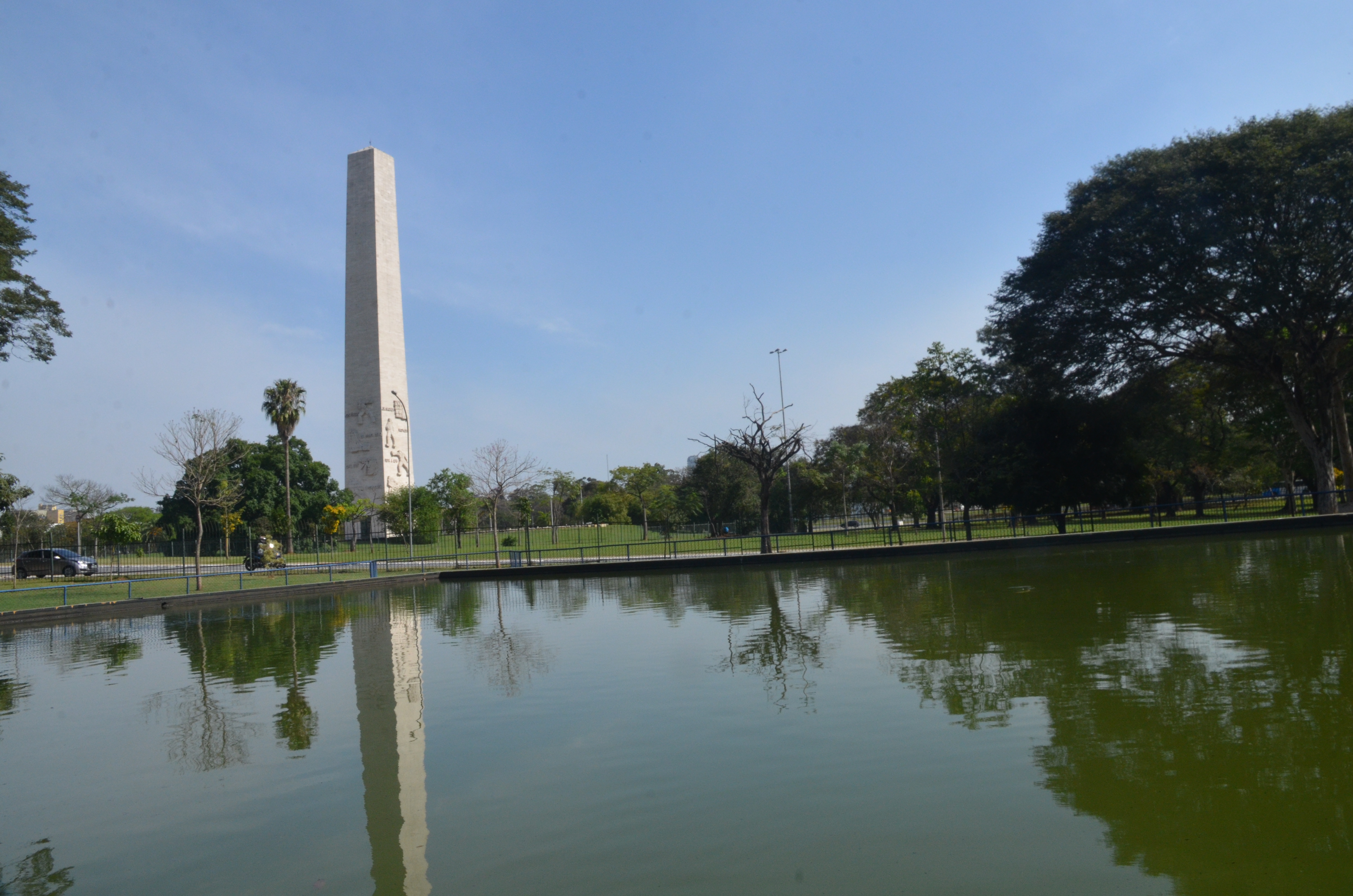 Na imagem um lago, muitas árvores e um carro localizados no Modelódromo do Ibirapuera, e logo atrás o monumento Obelisco.