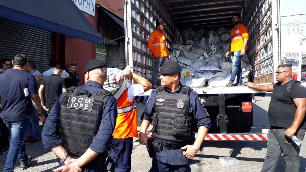Guardas Civis Metropolitanos, ao fundo um caminhão com produtos apreendidos e, homens de jaleco laranja.