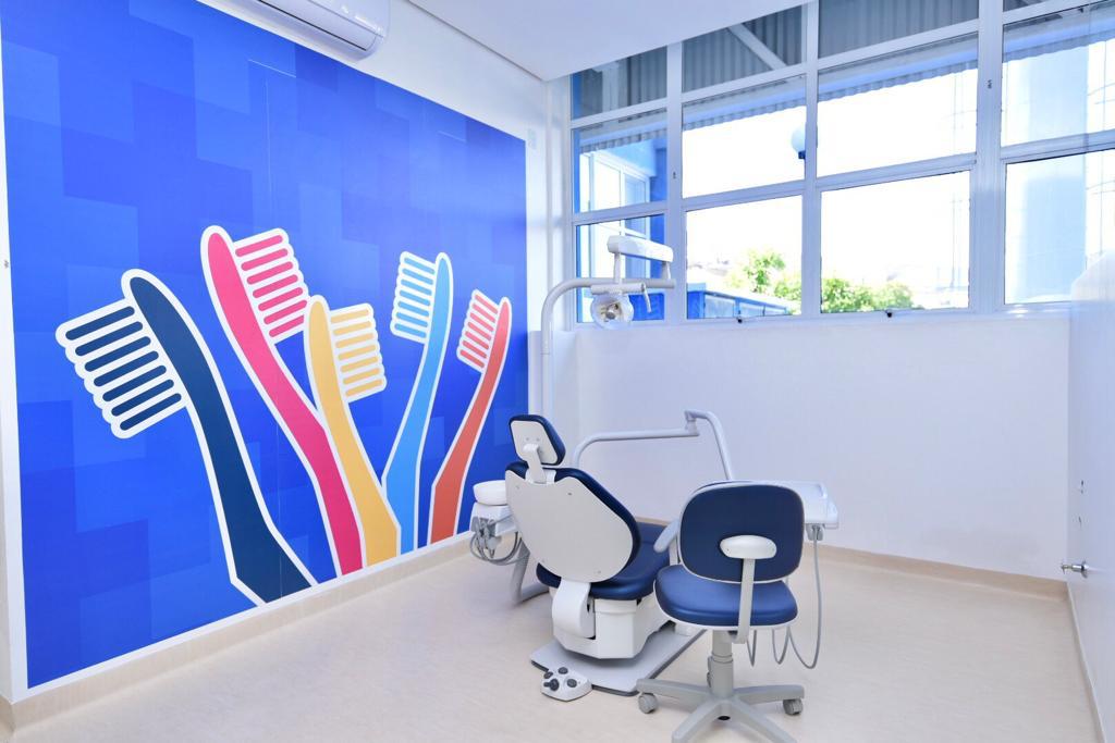 Na imagem há um consultório odontológico com equipamento e cadeira. Há também uma parede azul com várias escovas de dentes coloridas desenhadas.