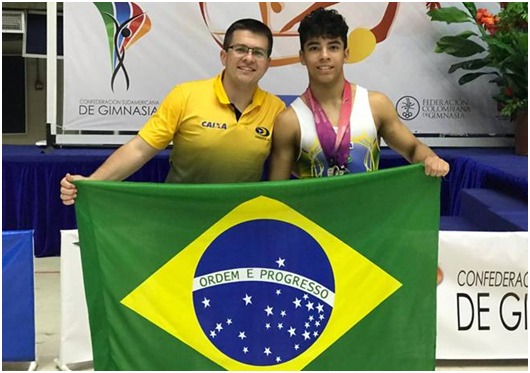 O atleta de ginástica artística Patrick Sampaio com uma medalha no pescoço junto com um integrante da comissão técnica, ambos segurando uma bandeira do Brasil.