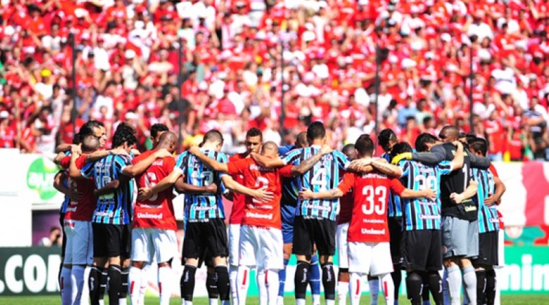 Na imagem, jogadores do Internacional e do Grêmio se abraçando formando um circulo no meio do campo, e atrás a torcida do Internacinal. Foto tirada em um dia ensolarado.