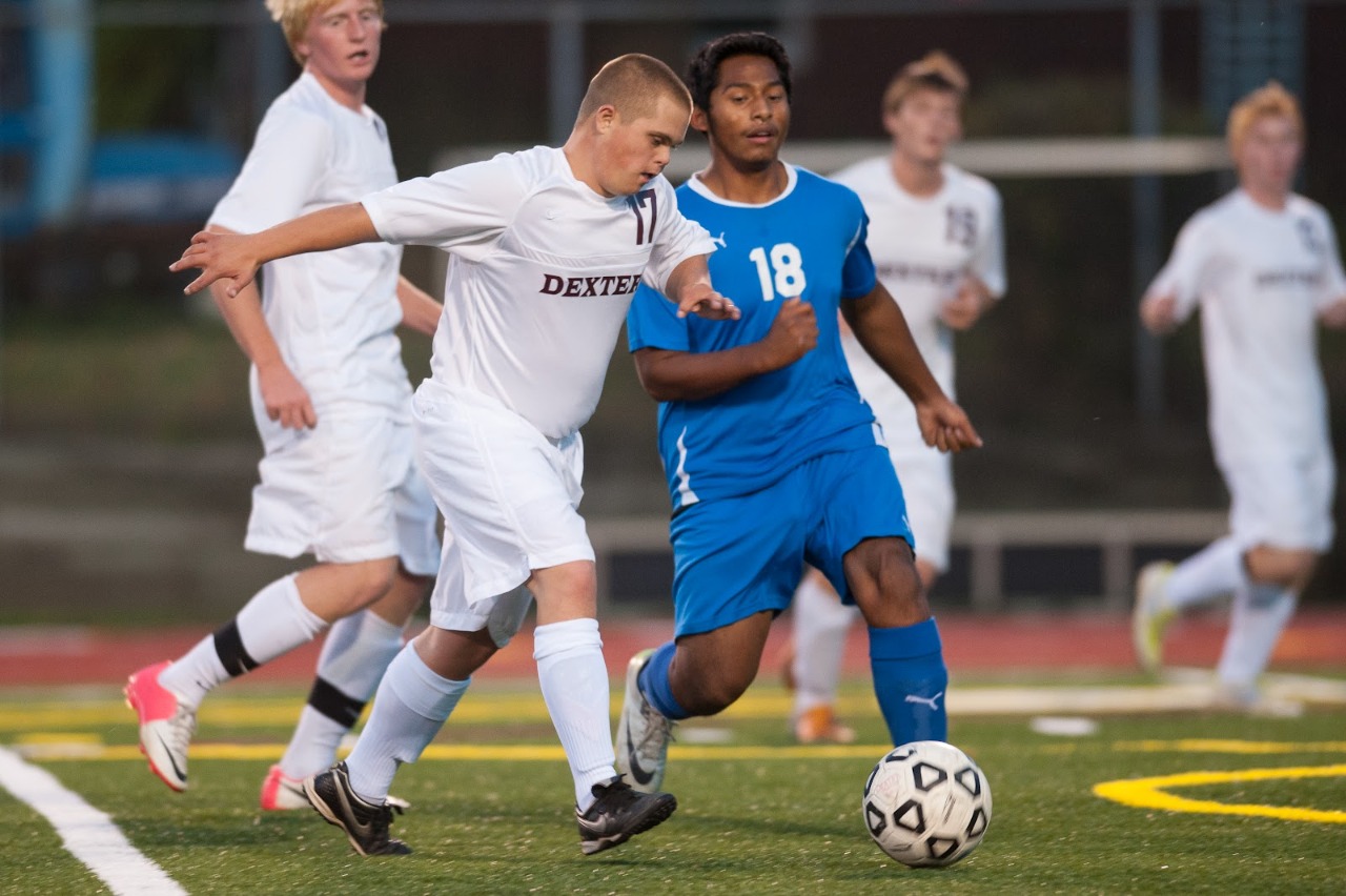 Atletas disputam a bola durante uma partida de futebol. Em destaque, um rapaz de branco e um rapaz de azul. Ao fundo, vários atletas de branco.