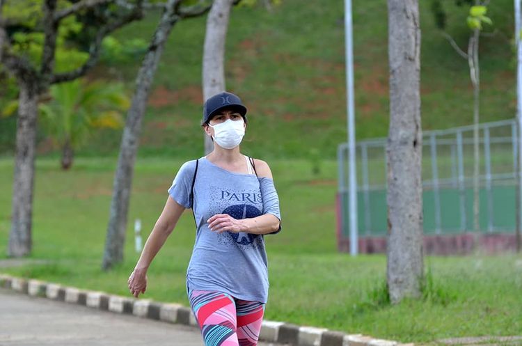 A imagem, tem uma mulher com boné e máscara, caminhando em um parque.