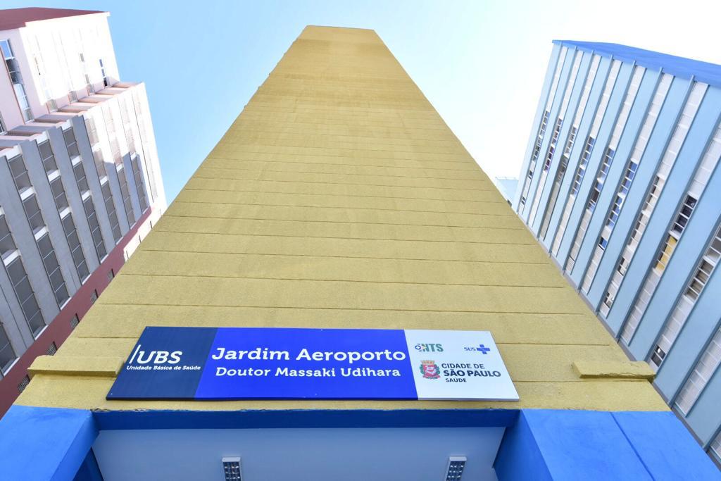 Fachada de prédio. Imagem tirada de baixo para cima. no centro há um prédio amarelo com a placa azul UBS Jardim Aeroporto. Há também um prédio de cada lado.