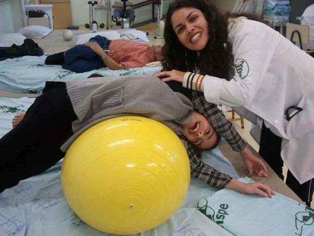 Na imagem, uma profissional de saúde usando jaleco branco está com uma mão em cima de uma menina que está deitada, apoiada em uma bola amarela. Atrás delas, está uma outra pessoa deitada em uma cama.