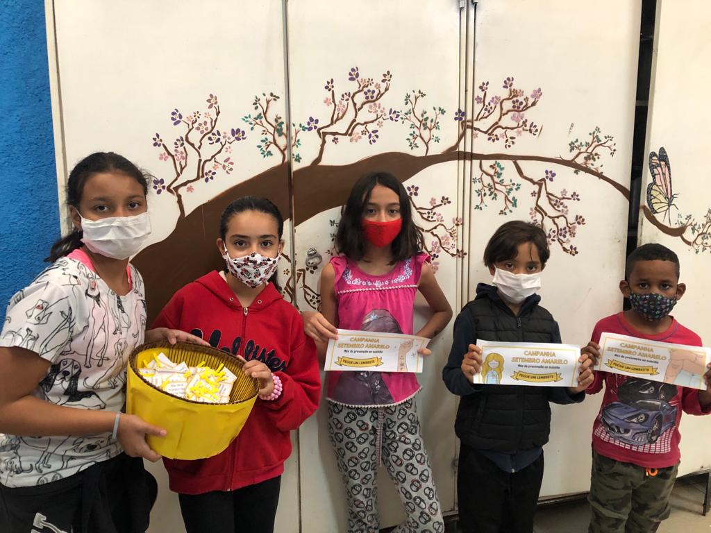 Cinco crianças participantes da atividade, todas com máscara, posam para a foto com uma cesta cheia dos bilhetes motivacionais escritos e faixas da campanha “Setembro Amarelo”.