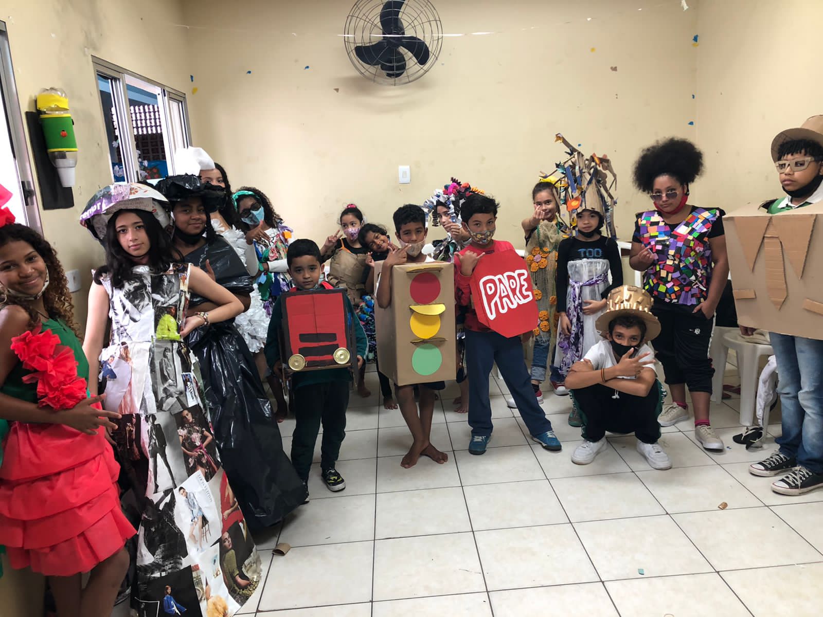Cerca de 16 crianças participantes do desfile, todas vestidas com fantasias diferenciadas produzidas com caixa, papéis, jornais, entre outros materiais, posam para foto nos bastidores da festa. 