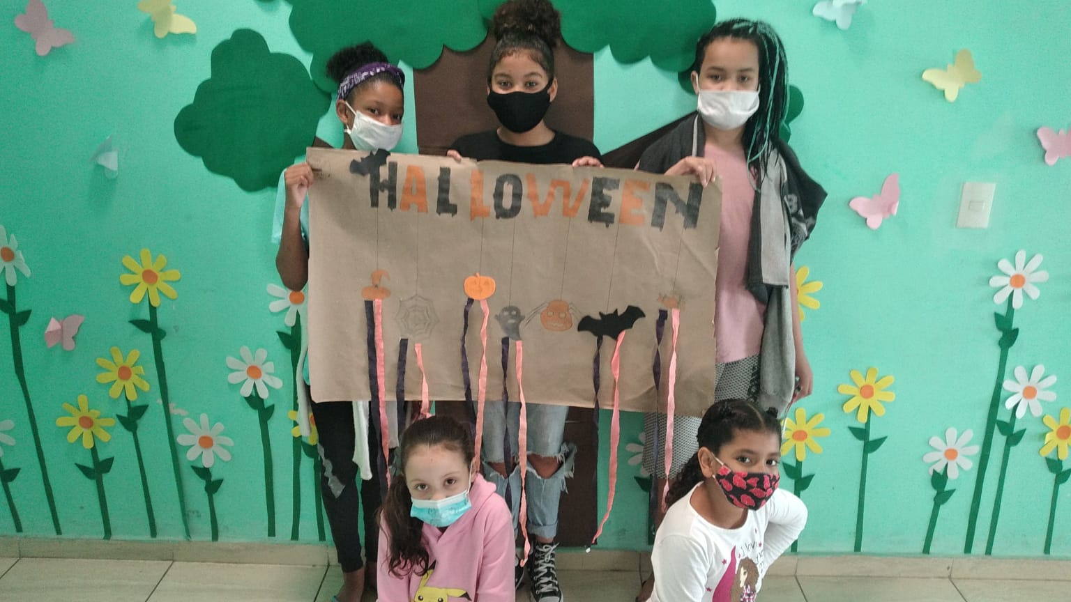 Quatro meninas posam para a foto com um dos cartazes preparados pelas próprias crianças para exposição na festa. No cartaz, escreveram a palavra “Halloween”, o Dia das Bruxas em inglês, em laranja e preto, com algumas decorações do tema.