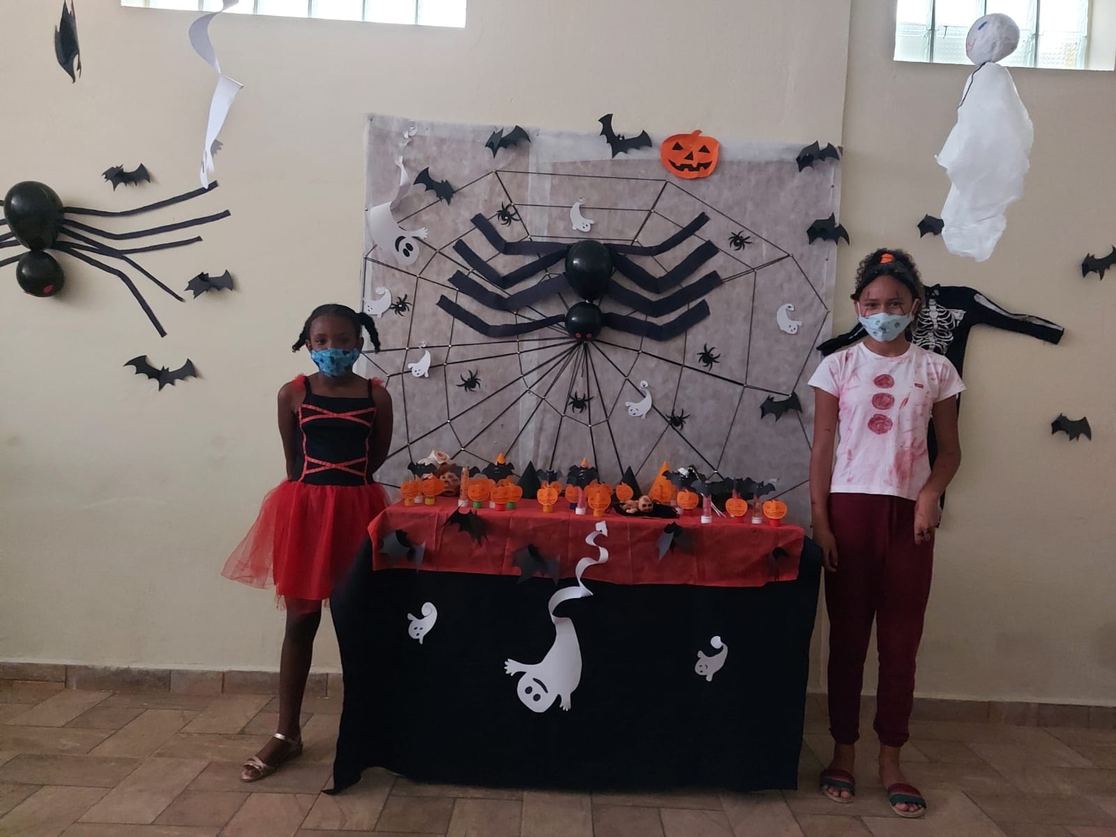 Duas crianças posam para foto com a mesa e o mural, decorados com apetrechos comemorativos e temáticos do Dia das Bruxas, como fantasmas, aranhas, abóboras e morcegos.