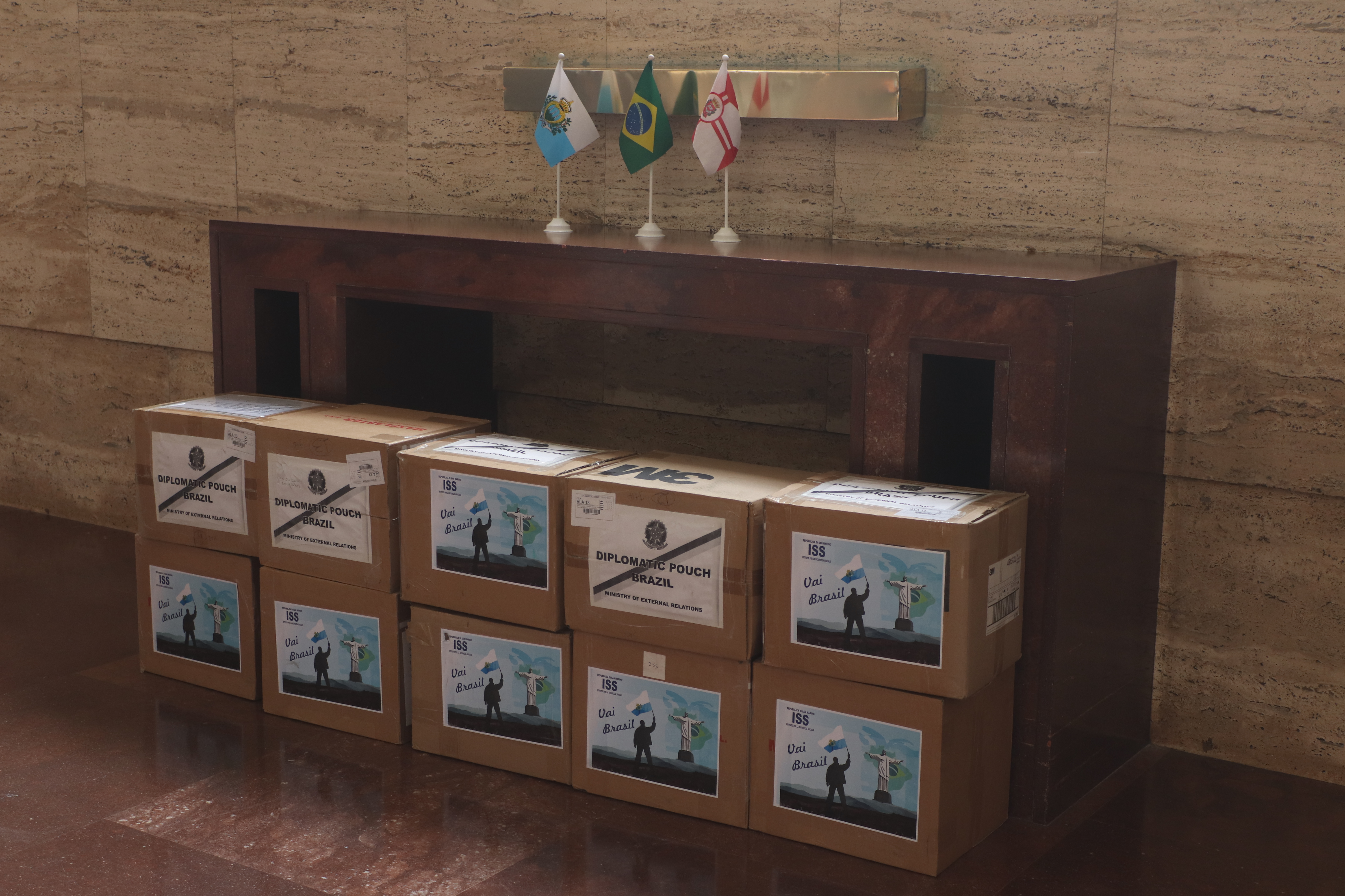 Foto das caixas com os equipamentos médico-hospitalares doados por San Marino, com as bandeiras de San Marino e da cidade de São Paulo colocadas em cima. 