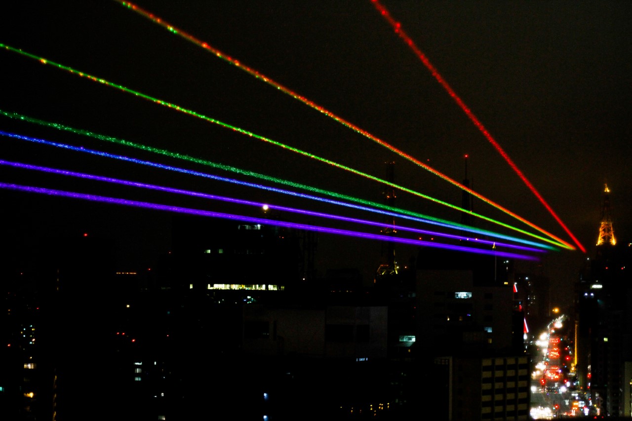 Foto de uma ação para o Dia da Visibilidade LGBTI no qual luzes com as cores da bandeira LGBT iluminam o céu da cidade de São Paulo durante a noite. 