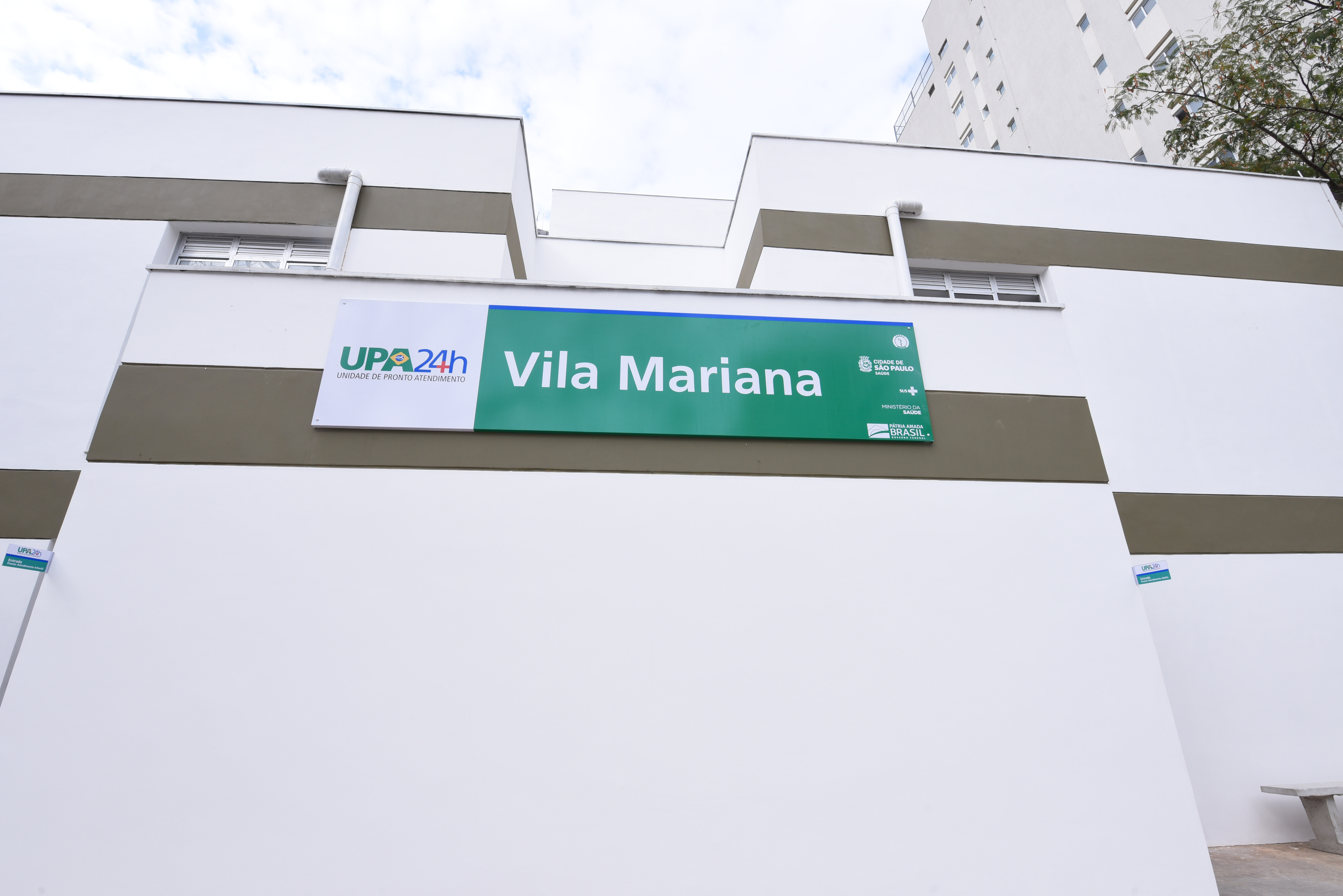 Foto de fachada. No centro de uma parede branca com faixa marrom, está uma placa verde com letras brancas "UPA Vila Mariana"