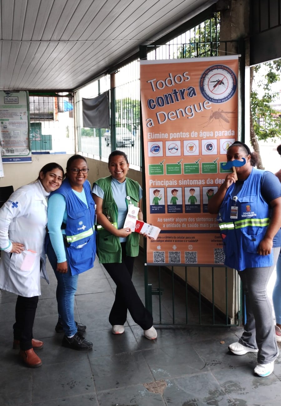 A foto mostra quatro agentes de saúde, meio ambiente e uma profissional de enfermagem junto a um banner ilustrado onde se lê “todos contra a dengue” e dicas ilustradas