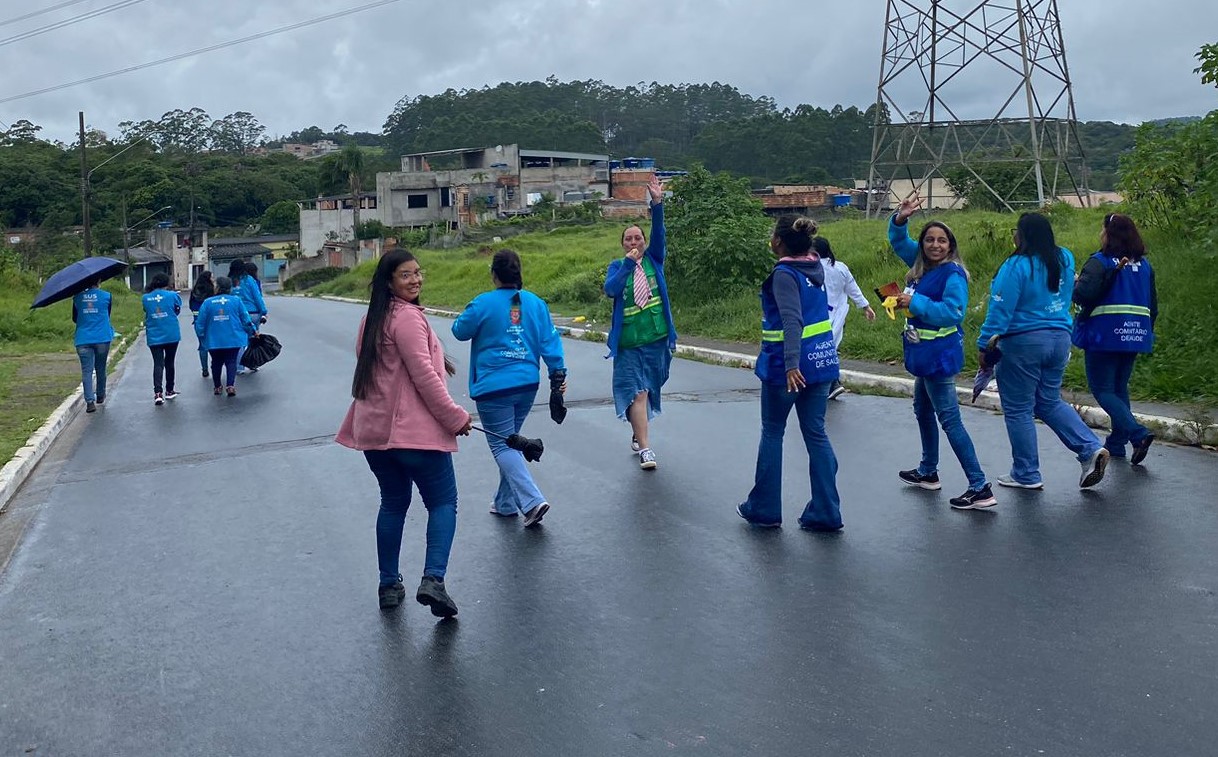 A foto, feita de trás, mostra mais de dez agentes de saúde caminhando em uma estrada de asfalto molhada pela chuva; ao fundo, vê-se uma comunidade, para a qual as agentes se encaminham