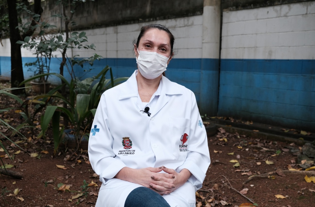Alessandra Groterria é uma mulher branca, jovem, que usa máscara e um jaleco com a logomarca do SUS e o símbolo da Prefeitura Municipal de São Paulo; ela está sentada em uma área ao ar livre, tendo ao fundo um muro pintado em branco e azul