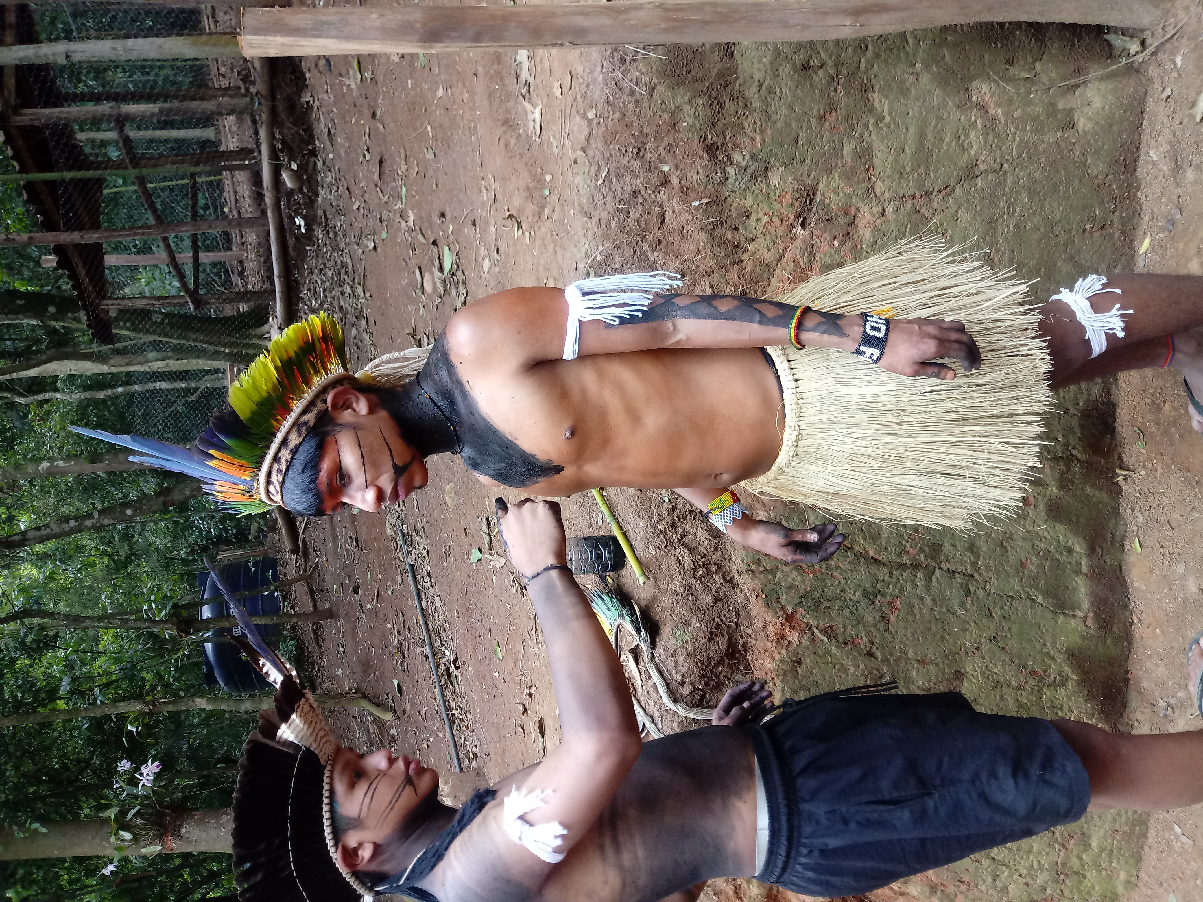 Na foto, vemos dois adolescentes indígenas; ambos usam cocares de penas e estão com os rostos pintados; um deles, que veste um calção, está pintando o torso do outro, que usa um saiote de palha, com tinta preta