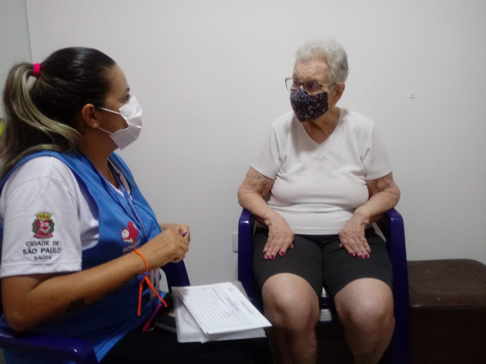 A foto mostra Gizelly, uma mulher jovem com uniforme da Secretaria Municipal da Saúde, interagindo com dona Miriam, uma mulher idosa; ambas estão de máscara e parecem conversar