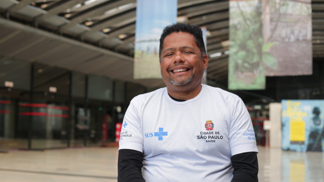 Edson do Nascimento Gomes é um homem negro de meia idade, que aparece sorrindo na foto; ele veste uma camiseta branca com a logomarca do SUS e o símbolo da cidade de São Paulo