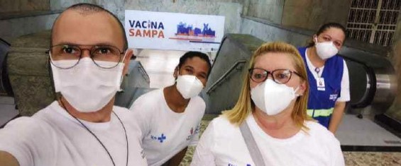 Eliezka está no centro da foto, atrás de duas pessoas e   ao lado de uma terceira. Eles estão em frente a uma   escada dentro de uma estação do metrô. Todos usam máscara   branca e camiseta branca com o emblema do SUS