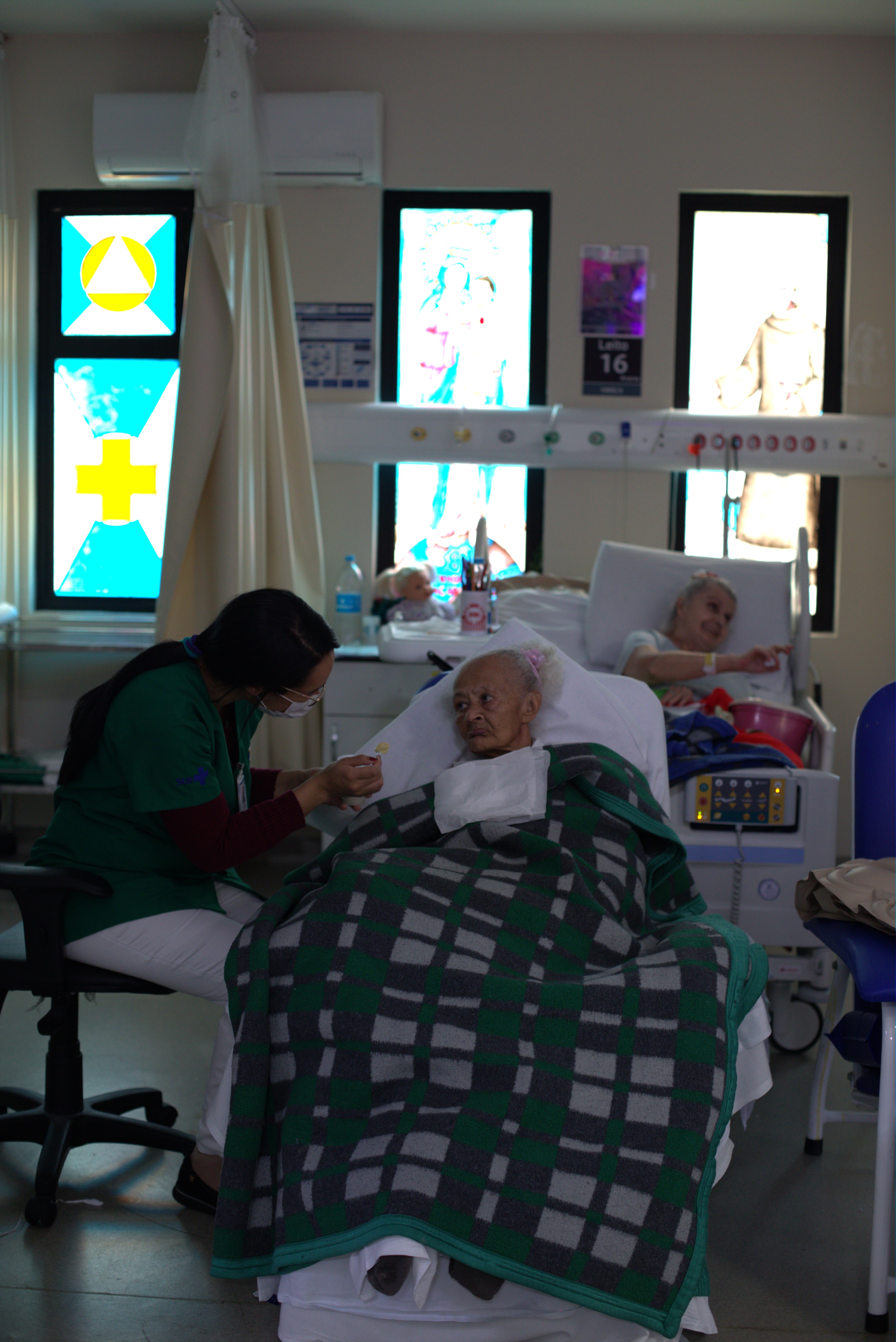A foto mostra uma enfermaria de hospital com destaque para duas camas; naquele que aparece em primeiro plano, uma mulher idosa recostada na cama e com um cobertor sobre seu corpo é alimentada na boca por uma profissional de enfermagem; em segundo plano aparece outra mulher, recostada na cama. A parede ao fundo é adornada com vitrais coloridos 