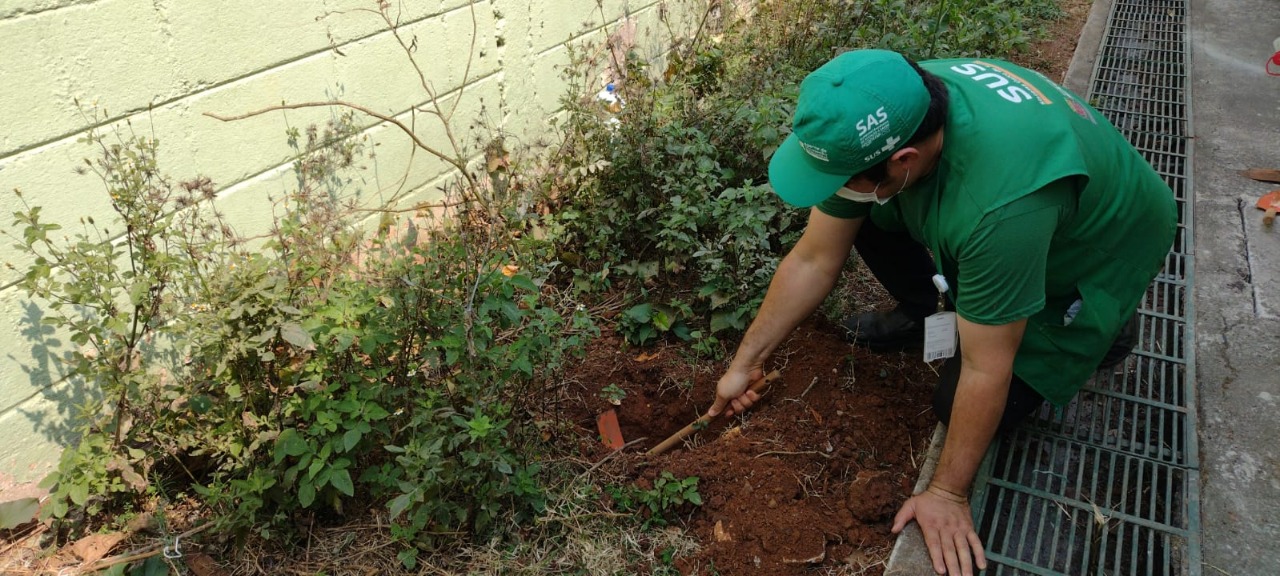 A foto mostra Everton agachado, cavando um buraco no solo da horta comunitária na UBS Carlos Muniz. Everton está de boné, colete e camiseta verde. Além disso, usa uma máscara facial branca. 