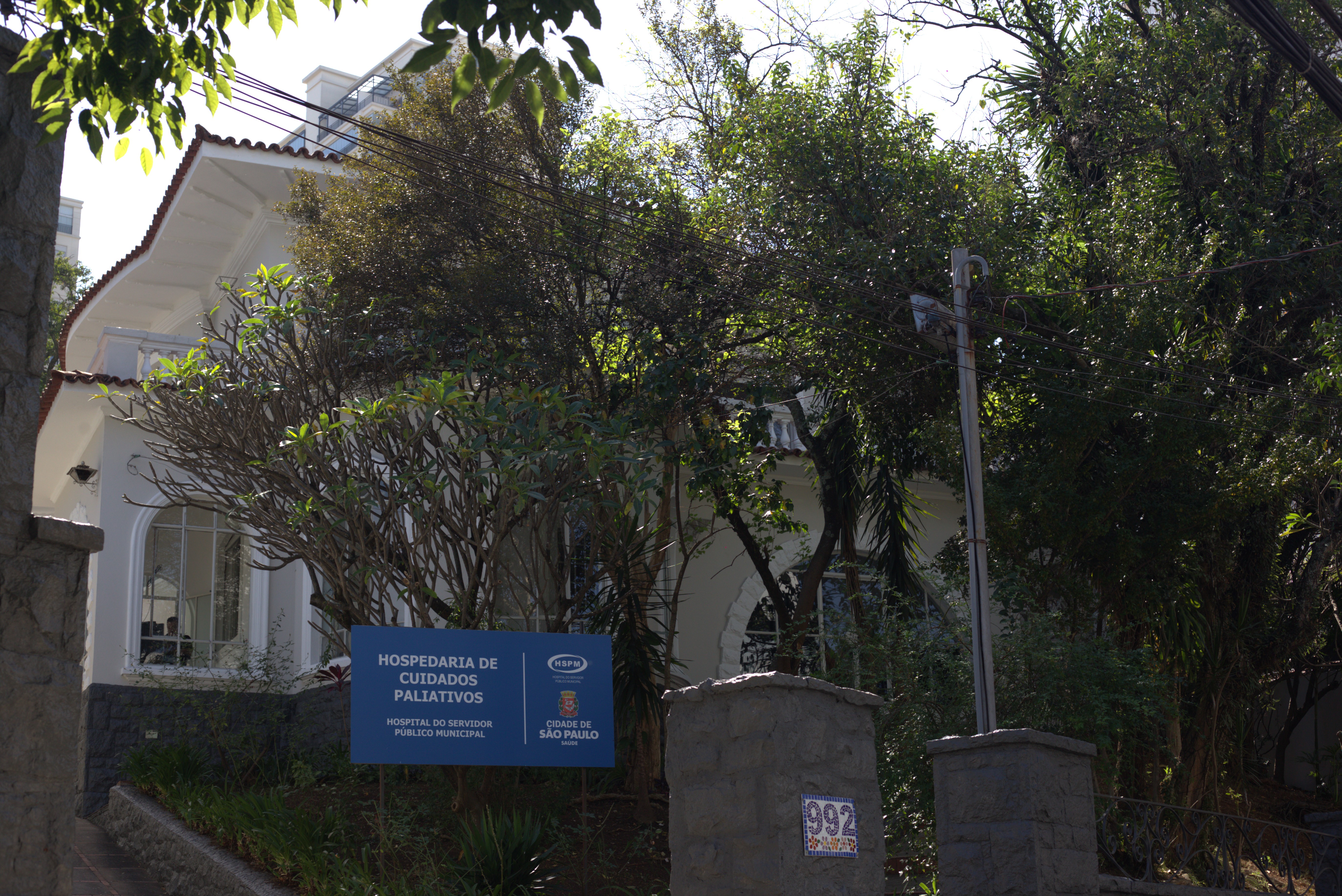A foto mostra a fachada de um casarão, pintado de amarelo claro e rodeado de árvores; em frente há uma placa azul, onde se lê “Hospedaria de Cuidados Paleativos – Hospital do Servidor Público Municipal”