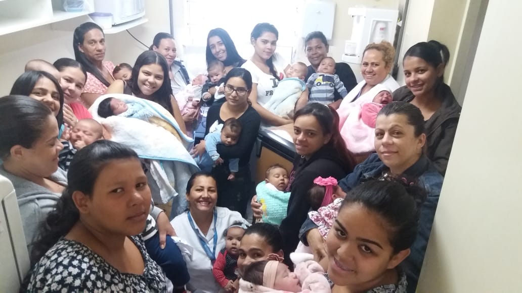 A foto mostra um grupo de cerca de 12 mulheres jovens, todas segurando bebês no colo; elas bem juntas, em um cômodo pequeno; no meio, está uma profissional de saúde, e em um dos cantos da sala vê-se um freezer de pequeno porte