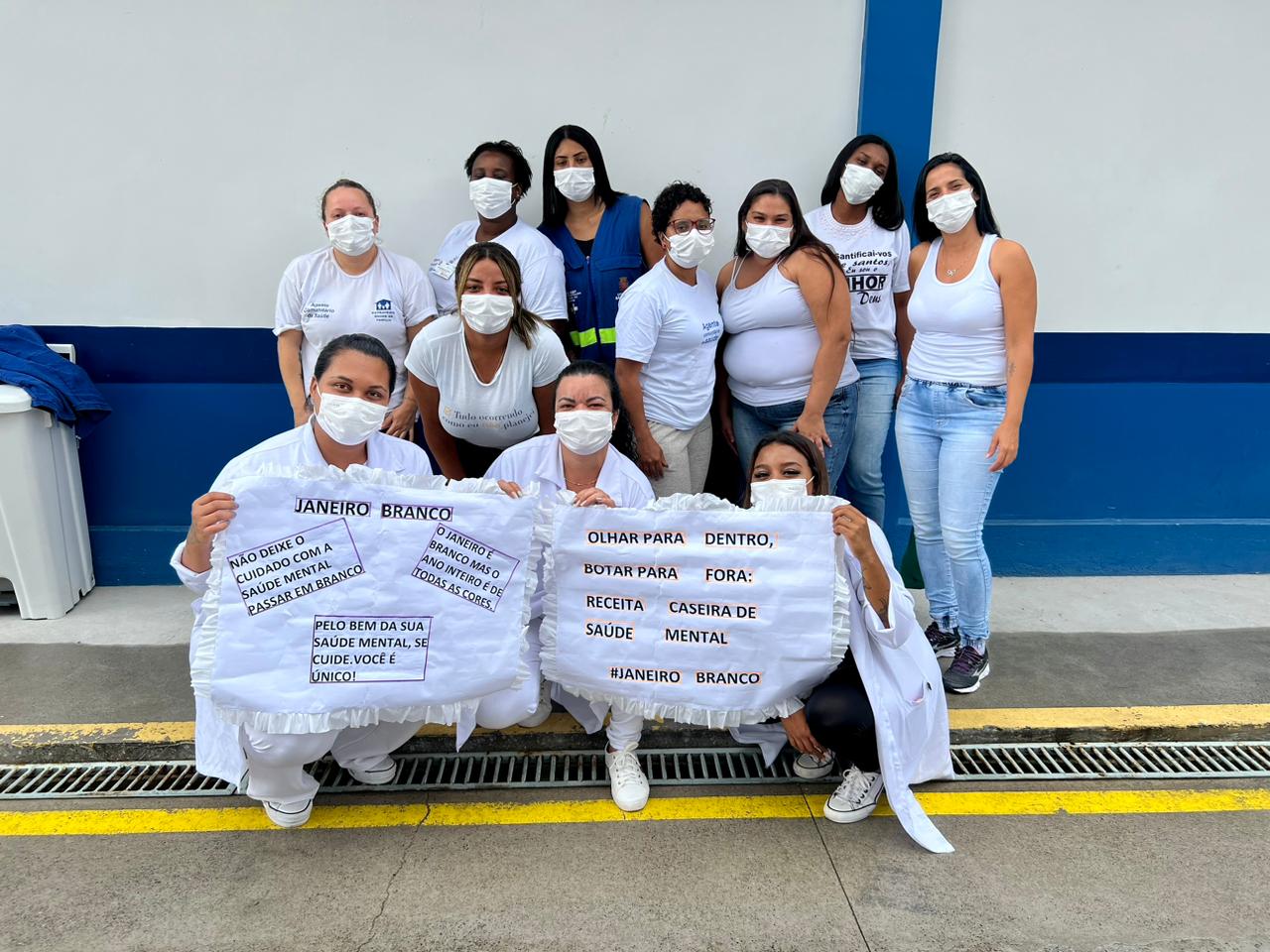 A foto mostra um grupo de 11 mulheres; elas estão em sua maioria de branco e usando máscaras, e algumas seguram cartazes com frases como "Não deixe o cuidado com a saúde mental passar em branco"