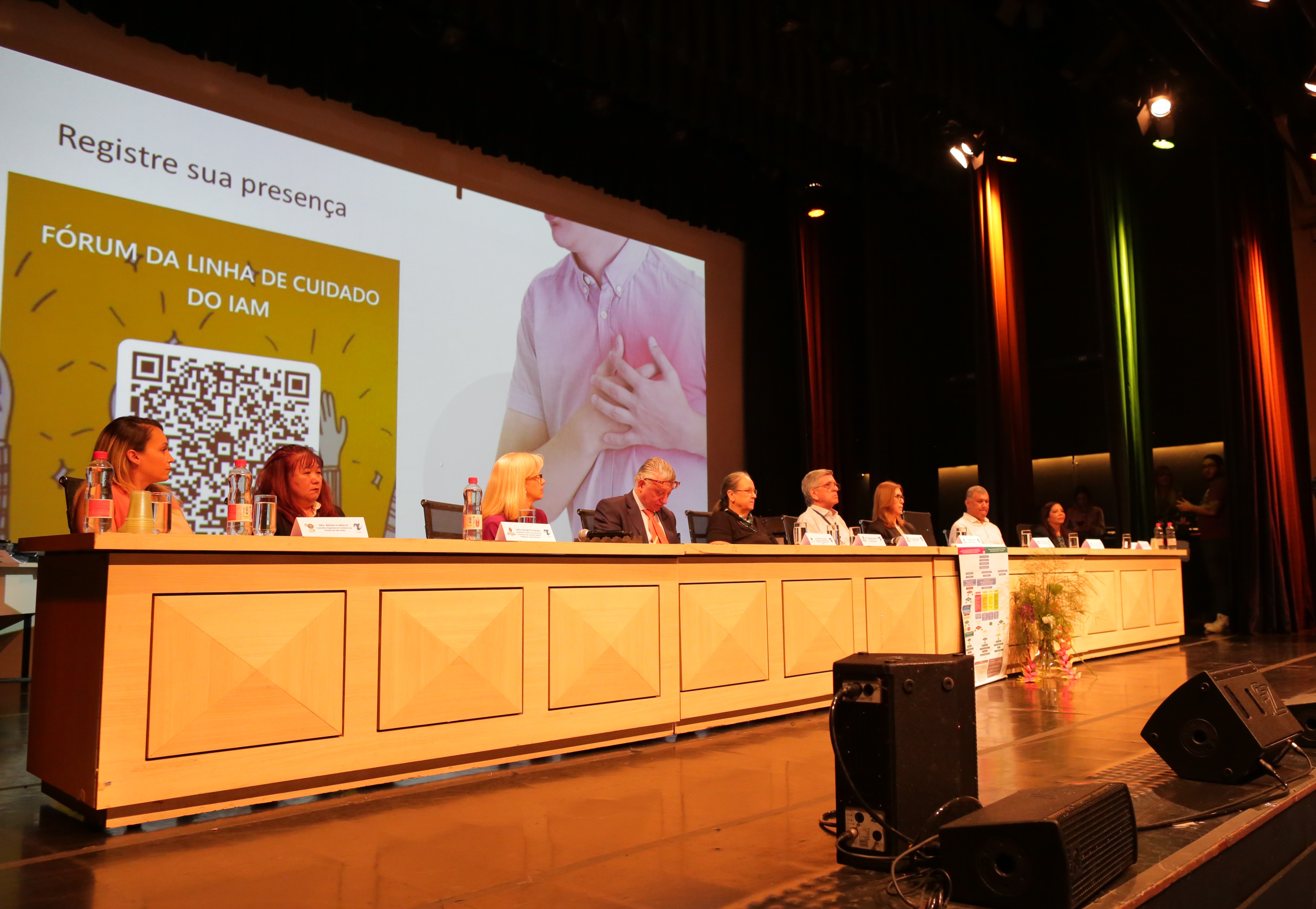 A foto mostra uma mesa em um evento, composta por vários homens e mulheres (ao fundo, vê-se um painel onde está escrito "Fórum da Linha de Cuidados do IAM