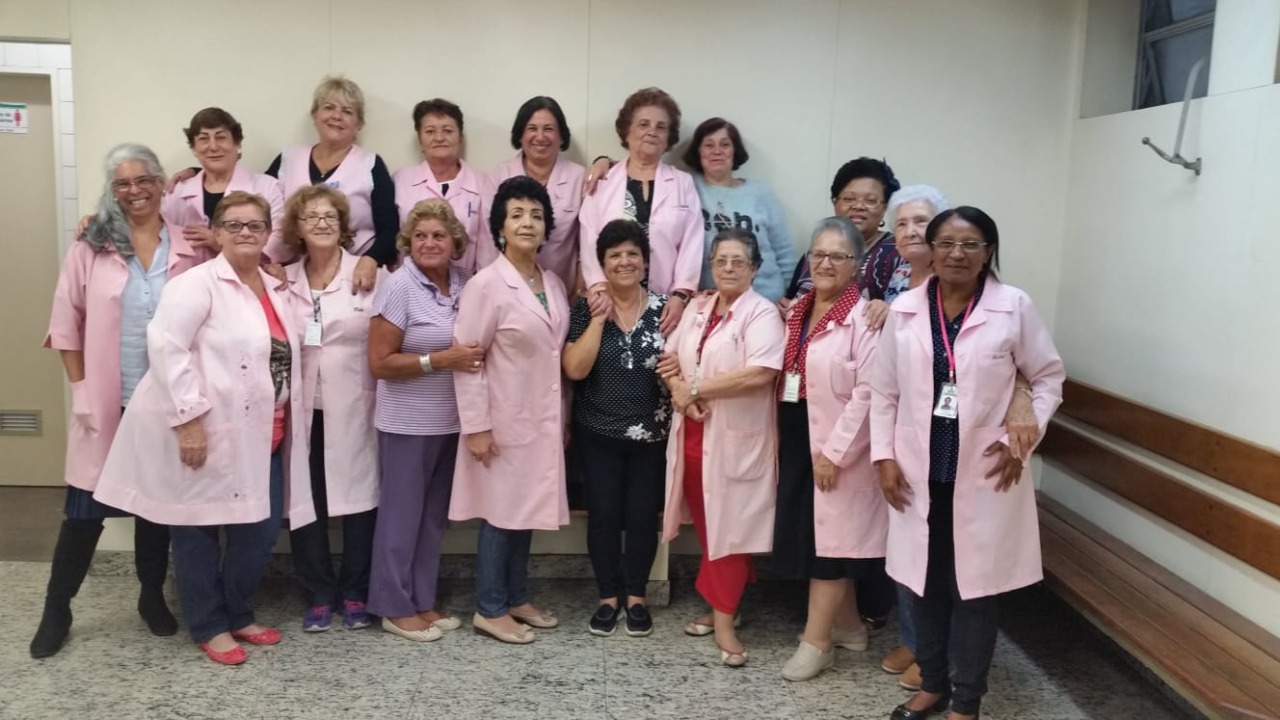 A foto mostra Marineusa, uma mulher de meia idade de pele negra, junto a um grupo de cerca de 15 mulheres, também de meia idade; elas usam um jaleco cor-de-rosa, que distingue os membros do programa de voluntariado 