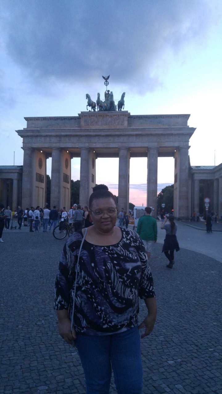 A foto mostra Marineusa posando em frente a um grande monumento, o Portão de Brandemburgo, na cidade de Berlim, na Alemanha; o monumento é composto por seis colunas e encimado por uma estátua mostrando uma deusa grega em uma biga puxada por quatro cavalos