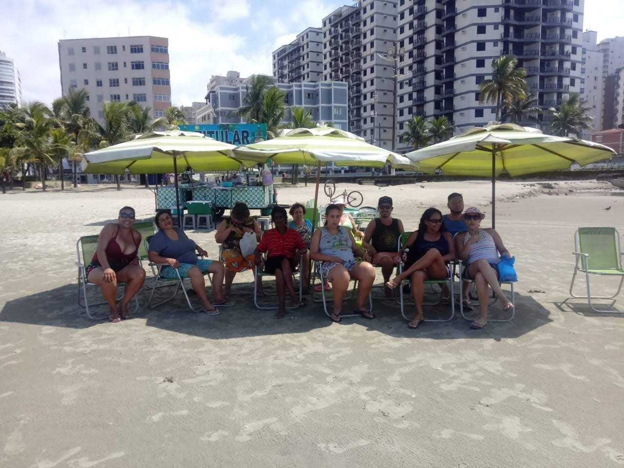A foto mostra as moradoras da Residência Terapêutica Mandaqui II sentadas sob guarda-sóis em cadeiras de praia, com os pés na areia  