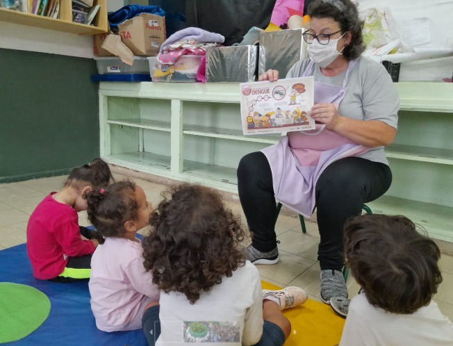 A foto mostra uma professora conversando com crianças pequenas, que estão sentadas à sua frente; a professora segura um pequeno cartaz ilustrado, onde se lê a palavra "dengue"