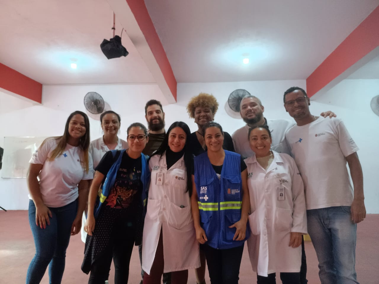 A foto mostra um grupo com dez pessoas, homens e mulheres; eles estão posando e sorriem; todos usam jaleco e camisetas com logomarca do SUS e brasão da Prefeitura de São Paulo  