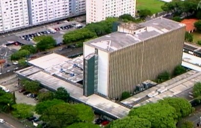 A foto mostra a vista aérea de um prédio de dez andares; a estrutura está cercada por árvores 
