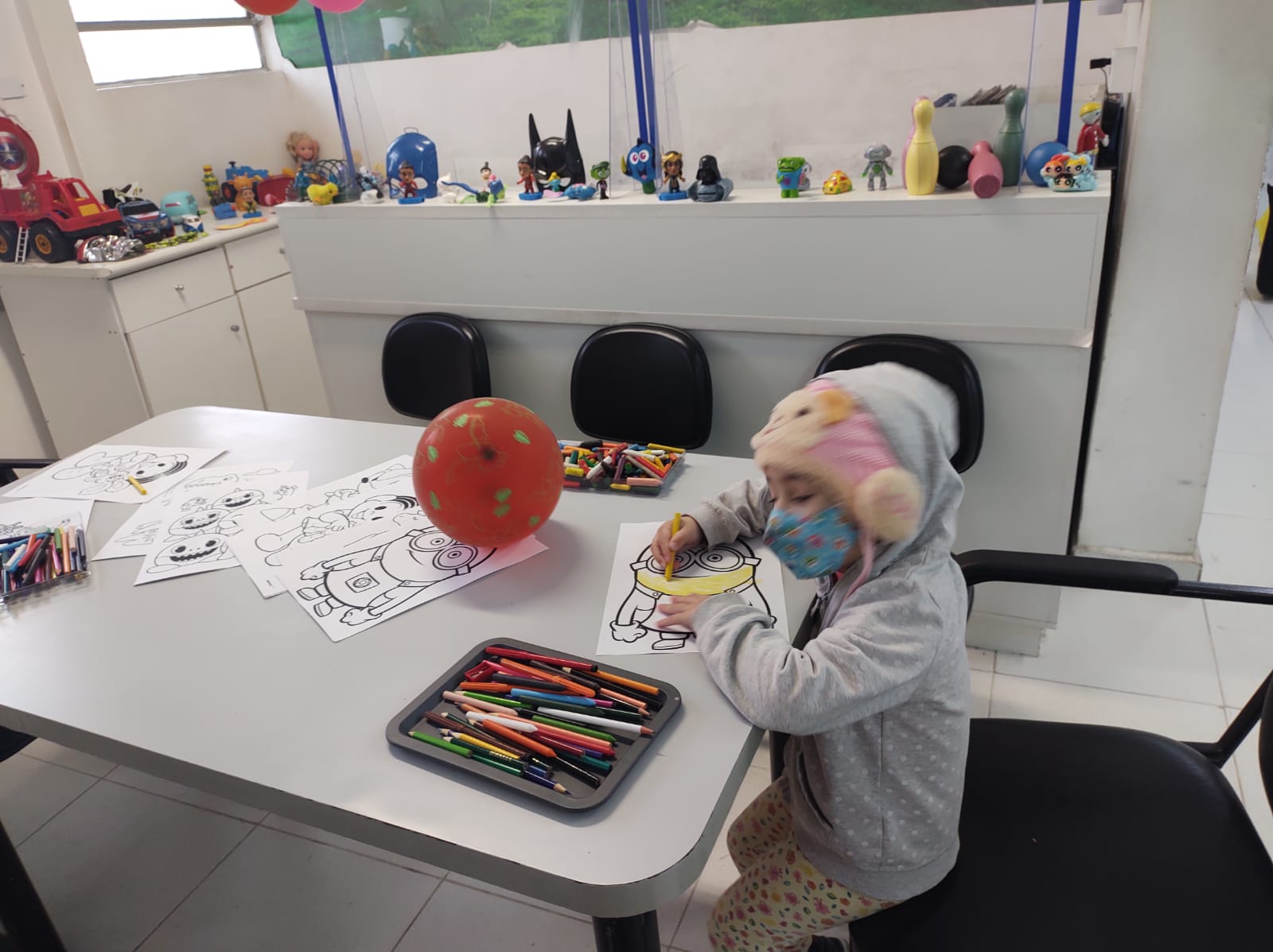 Foto mostra uma criança usando touca e máscara; ela está sentada junto a uma mesa onde estão várias folhas com desenhos além de lápis de cor, ela colore um dos desenhos
