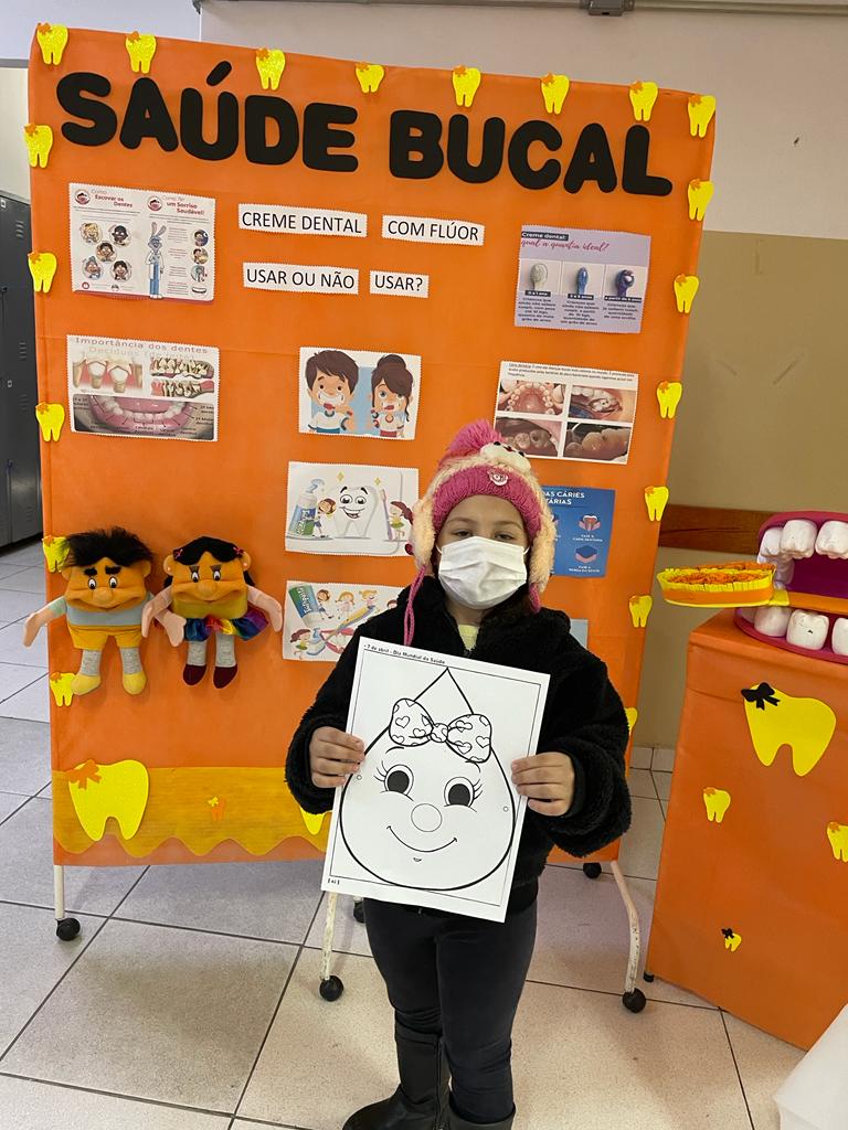 A foto mostra uma criança usando touca e máscara; ela segura uma folha com o desenho de uma gota, para colorir, e está em frente a um painel com textos e fotos sobre saúde bucal