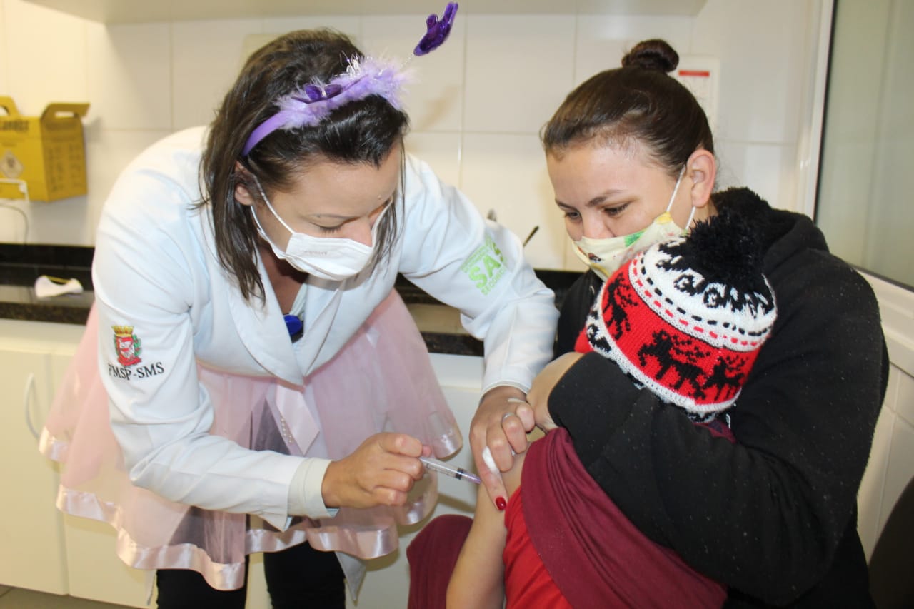 A foto mostra uma criança sentada no colo de uma mulher, enquanto recebe uma vacina injetável no braço esquerdo, aplicada por uma mulher que usa jaleco branco, tiara com antenas na cabeça e uma saia cor-de-rosa de bailarina por cima da roupa