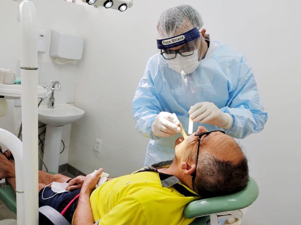 Na imagem há um dentista em pé vestindo roupa de proteção azul, máscara e touca branca. Ele está mexendo usando uma espátula para ver a boca de um homem que está deitado em uma maca. O homem usa camisa amarela e óculos.