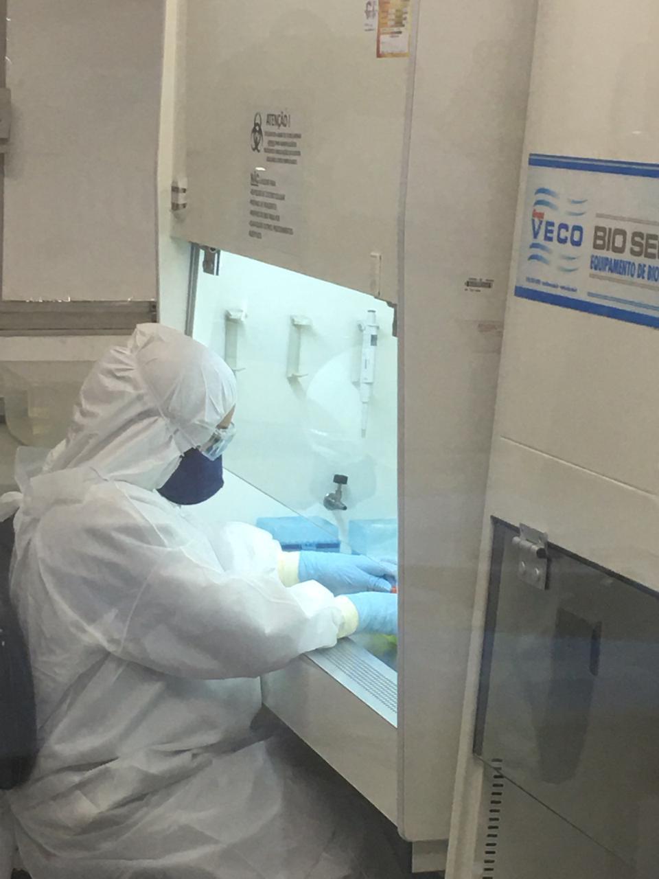 Imagem de uma profissional de saúde vestindo jaleco de proteção branco e máscara azul. Ela está em frente a uma máquina, com as mãos apoiadas em cima de uma estrutura.