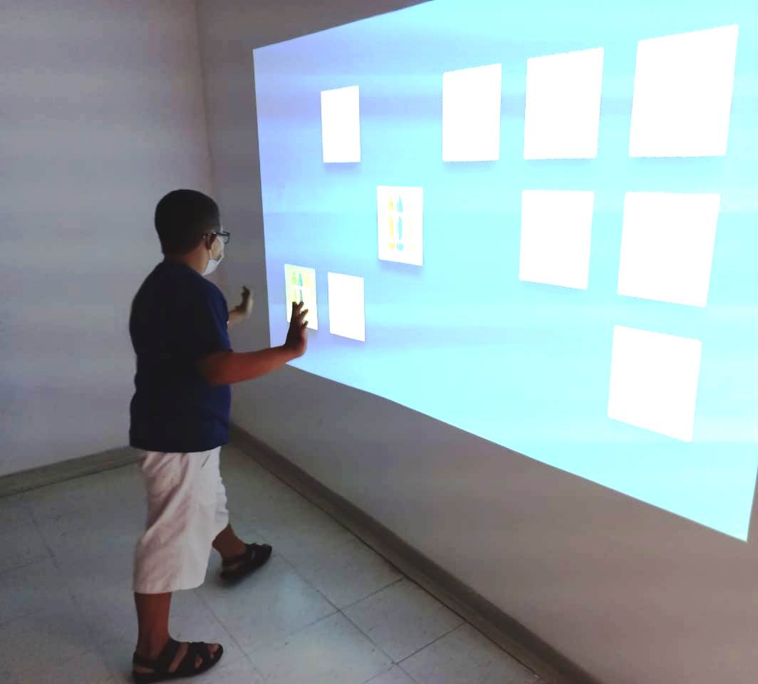 Um menino jovem está em frente a uma parede que tem uma tela projetada. Na tela azul, estão alguns quadrados brancos separados, como um jogo da memória.