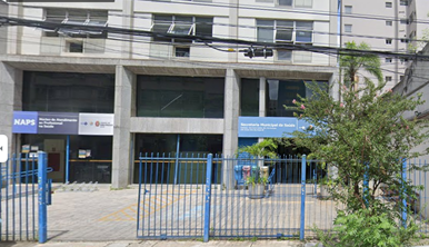 Na imagem está a entrada do prédio da Secretaria Municipal da Saúde. O prédio possui duas entradas. A entrada que está à esquerda é o NAPS, que possui uma placa  na qual está escrito NAPS.