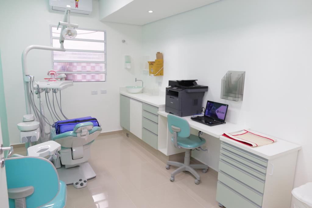 A foto mostra uma sala de atendimento odontológica equipada com cadeira, equipamentos e ar condicionado; ao longo de uma das paredes está um balcão com lavatório de um lado e notebook e impressora de outro