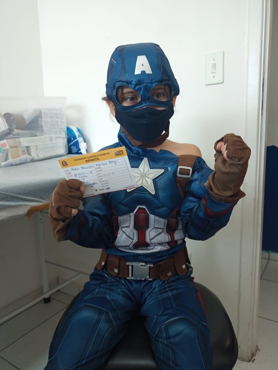 Foto de um menino de aproximadamente cinco anos sentado em uma cadeira. Ele está usando a fantasia de um super-herói azul, Capitão América. Atrás dele, está uma parede branca.