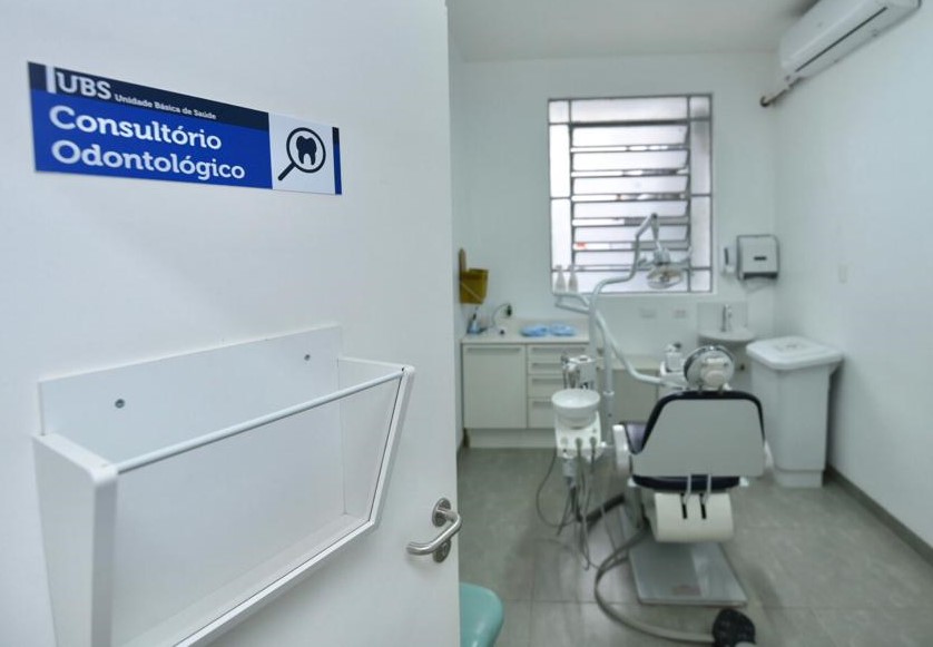 Foto de consultório odontológico. À esquerda da imagem, uma porta com uma placa azul com letras brancas diz "Consultório Odontológico". As paredes são brancas, com uma janela em uma delas e um ar condicionado em outra.