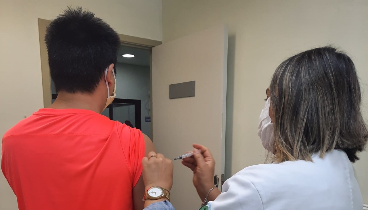 Foto de vacinação. À direita da imagem, uma profissional de saúde vestindo jaleco branco está aplicando a dose da vacina em um rapaz mais alto, que está ao seu lado, de costas. Ele veste uma camisa vermelha e está levando uma das mangas. 