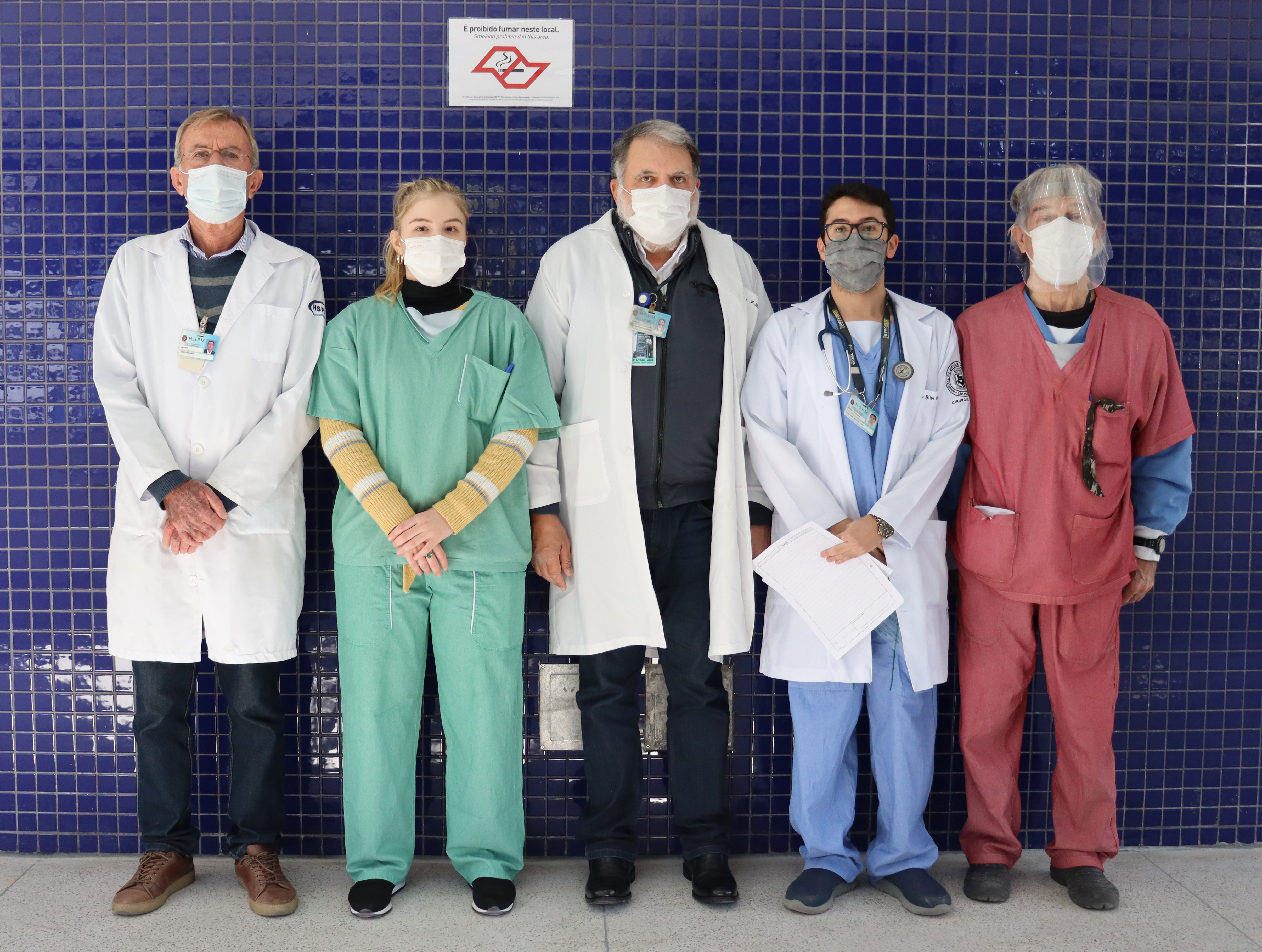 Foto de cinco cirurgiões do hospital em frente a uma parede de ladrilho azul escuiro.
