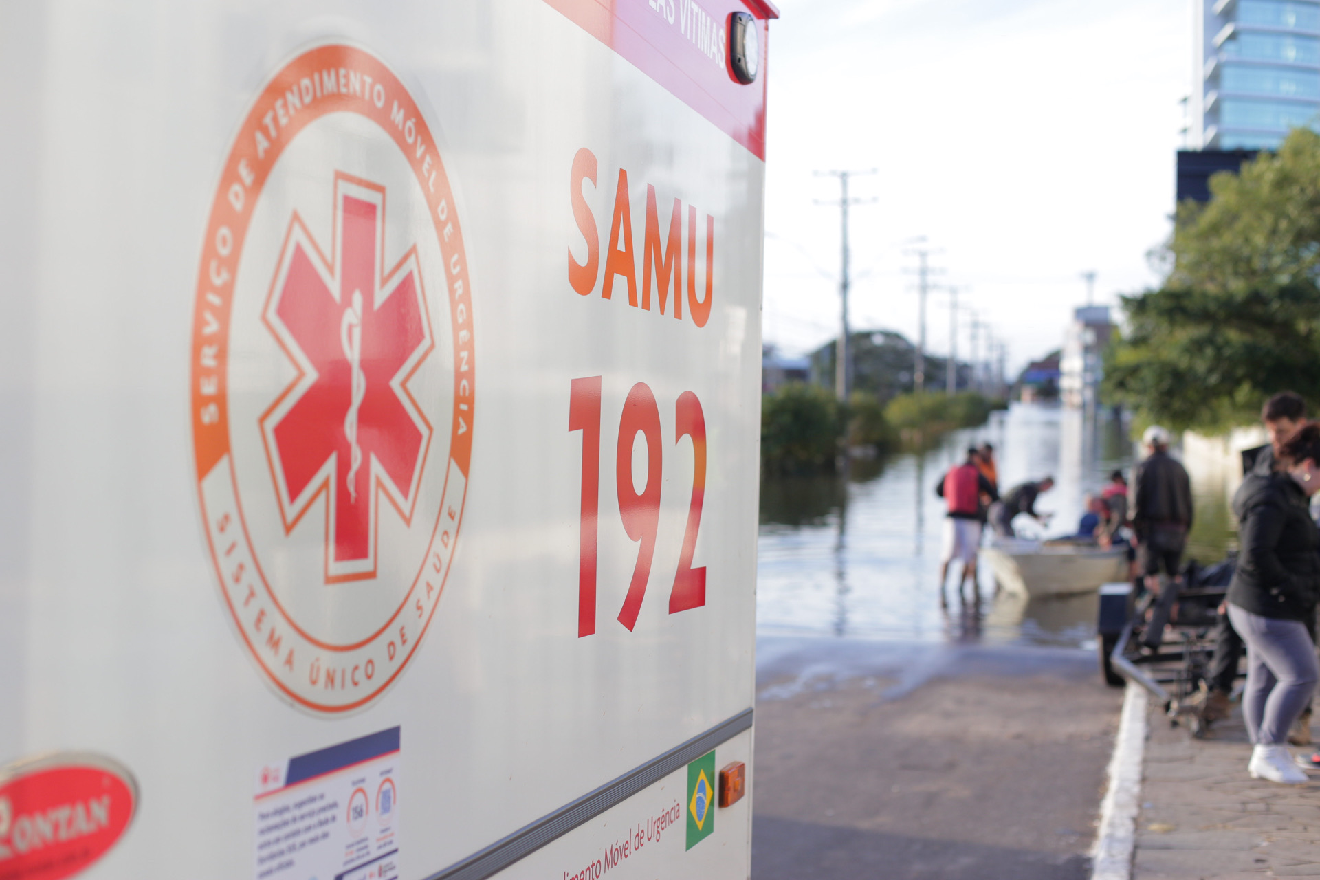 A foto mostra em primeiro plano a logomarca do Samu-192; ao fundo, em segundo plano, vê-se água cobrindo o asfalto da rua e pessoas em volta de uma canoa 