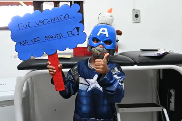 Um menino de aproximadamente oito anos está vestindo uma fantasia azul de Capitão América. Ele segura uma placa azul que tem letras pretas escrevendo a frase "Fui vacinado na UBS Santa Fé". O menino faz um sinal de positivo com uma das mãos.